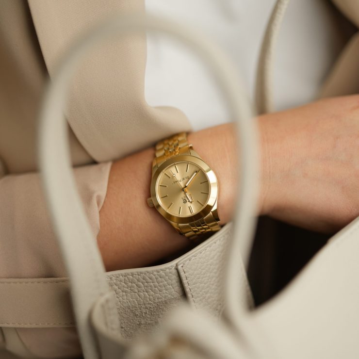 Goldfarbene Armbanduhr mit Gliederarmband am Handgelenk einer Person. Die Uhr wird in Kombination mit heller Kleidung und einer Handtasche getragen und unterstreicht einen stilvollen, eleganten Business-Look.