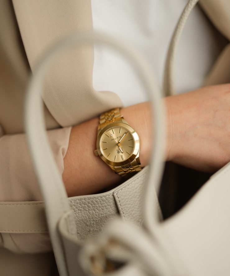 Goldfarbene Armbanduhr mit Gliederarmband am Handgelenk einer Person. Die Uhr wird in Kombination mit heller Kleidung und einer Handtasche getragen und unterstreicht einen stilvollen, eleganten Business-Look.