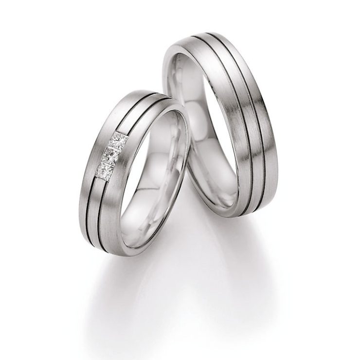 Zeitloses Trauring-Set in eleganter Silberoptik mit fein mattierter Oberfläche und dezenten, umlaufenden Zierlinien. Der Damenring wird durch drei quadratisch gefasste Diamanten stilvoll veredelt und setzt einen modernen, funkelnden Akzent. Die klare Formensprache und das harmonische Design machen diese Ringe zu einem besonderen Symbol für Liebe, Treue und Beständigkeit.