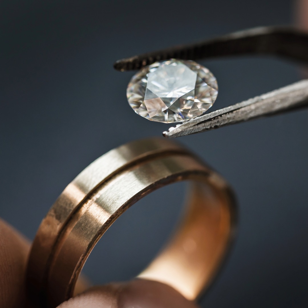 Pinzette hält einen funkelnden Diamanten über einem roségoldfarbenen Ring, bereit für die Einfassung in das Schmuckstück