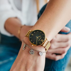 Nahaufnahme einer goldfarbenen Armbanduhr mit schwarzem Zifferblatt und Mesh-Armband, die zusammen mit einem silbernen Charm-Armband mit Herz- und Friedenssymbol-Anhängern getragen wird.