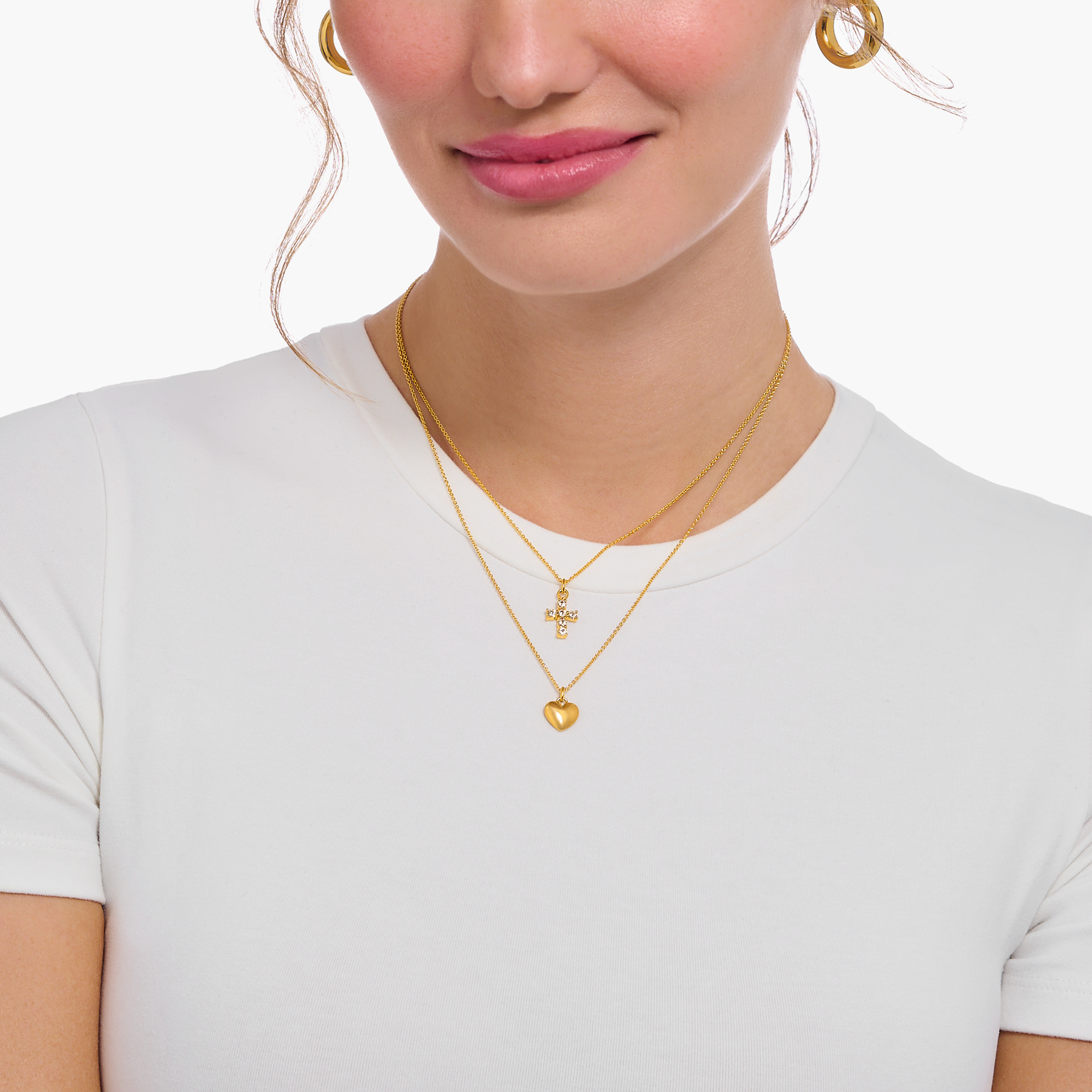 Model trägt zwei feine goldfarbene Halsketten im Layering-Stil mit einem Herzanhänger und einem funkelnden Kreuzanhänger, kombiniert mit goldfarbenen Creolen und weißem Oberteil