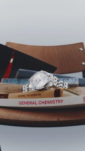 Tissot Powermatic 80 Automatikuhr aus Edelstahl auf einem Stapel von Lehrbüchern neben einem Abschluss-Hut auf einem Holzstuhl – Symbol für Studium, Erfolg und zeitlosen Stil.
