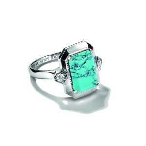 Ausdrucksstarker Ring aus poliertem Silber mit einem rechteckigen Schmuckstein in leuchtendem Türkisblau und charakteristischer dunkler Maserung. Der Stein ist in einer markanten geometrischen Fassung eingefasst und wird von kleinen funkelnden Ziersteinen auf beiden Seiten akzentuiert. Die breite, leicht geteilte Ringschiene unterstreicht das elegante und moderne Design. Präsentiert auf hellem Hintergrund mit dekorativem Schatten.