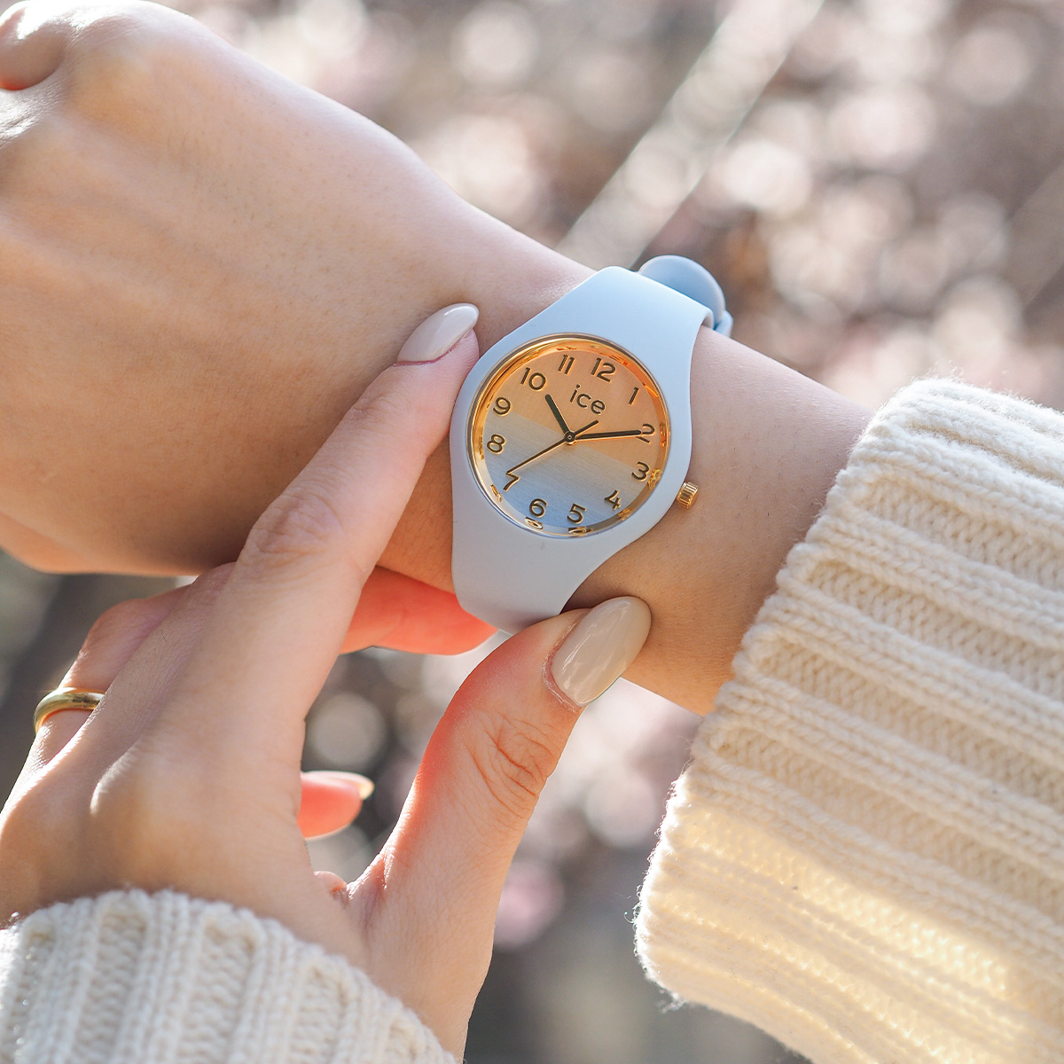 Person trägt eine ICE Watch mit hellblauem Silikonarmband und goldenem Zifferblatt am Handgelenk. Die Uhr wird im warmen Tageslicht getragen und unterstreicht einen modernen, eleganten Freizeitlook.