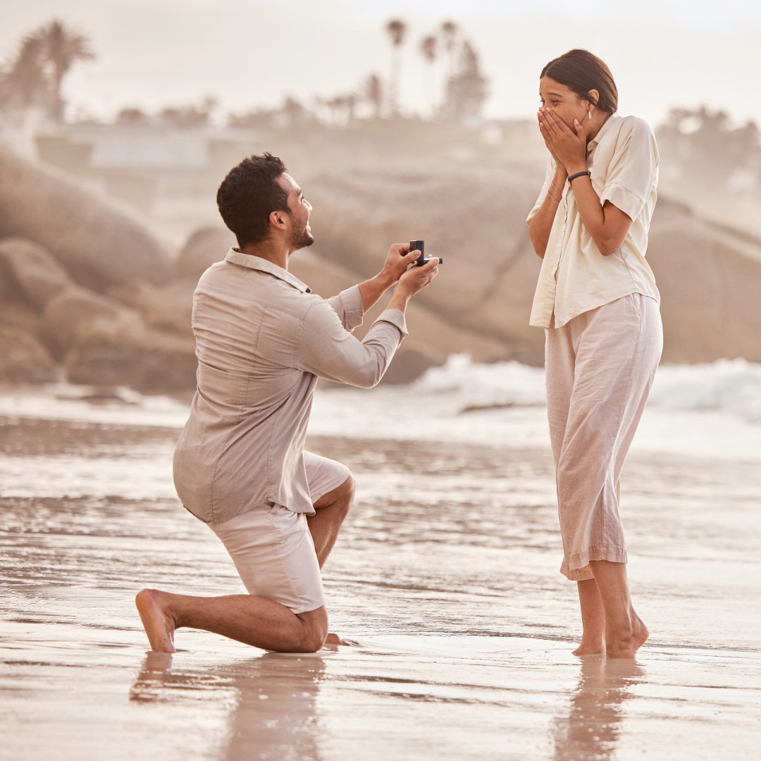 Emotionaler Moment eines romantischen Heiratsantrags am Meer: Ein Mann kniet am Strand und hält einen Verlobungsring bereit, während seine Partnerin überrascht und überwältigt reagiert. Die stimmungsvolle Kulisse bei Sonnenuntergang unterstreicht die Bedeutung dieses unvergesslichen Augenblicks voller Liebe und Zukunftsversprechen.
